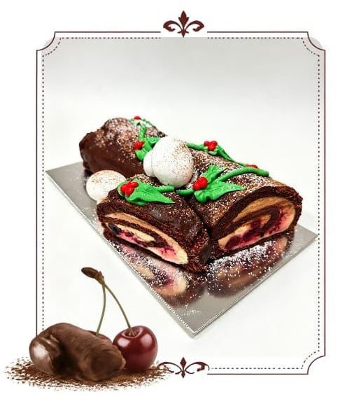 Chocolate Bûche de Noël cake decorated with holly, meringue, and powdered sugar, showing cherry and cream filling. Cherries a