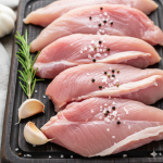 Turkey Buffs Raw turkey cutlets seasoned with salt and peppercorns, alongside fresh rosemary and garlic cloves on a dark cutting board.